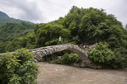 Georgia, Adjara, Makhuntseti, Young man crossing stone arch bridge stretching over brown river - WVF01687