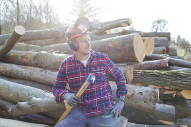 Nachdenklicher Holzfäller, der eine Axt hält, während er an einem sonnigen Tag auf einem Baumstamm sitzt - HMEF00939