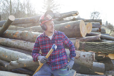 Thoughtful lumberjack holding axe while sitting on logs during sunny day - HMEF00939