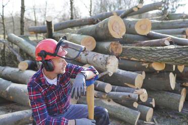 Nachdenklicher Holzfäller, der eine Axt hält, während er neben Baumstämmen im Wald sitzt - HMEF00940