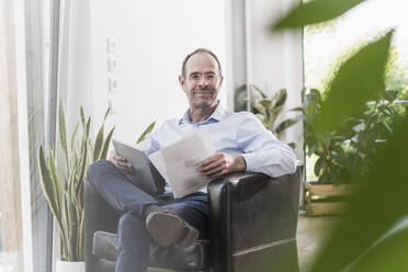 Portrait of smiling businessman with digital tablet and documents working at home - UUF20374