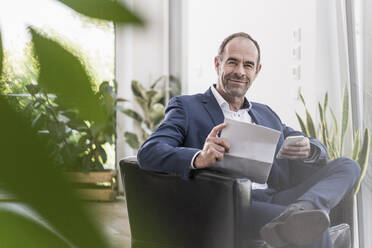 Portrait of smiling businessman with smartphone and documents working at home - UUF20376