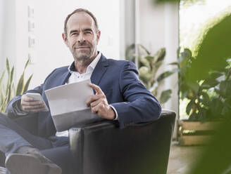 Portrait of smiling mature businessman with smartphone and documents working at home - UUF20377