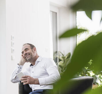 Porträt eines lächelnden reifen Mannes mit Smartphone im Sessel sitzend - UUF20383