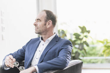 Portrait of pensive businessman sitting in armchair looking at distance - UUF20386