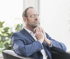 Portrait of businessman wearing glasses sitting in armchair meditating with eyes closed - UUF20392