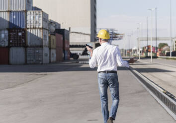 Rückenansicht eines Geschäftsmannes mit Schutzhelm, der auf einem Industriegelände ein Mobiltelefon benutzt - UUF20406