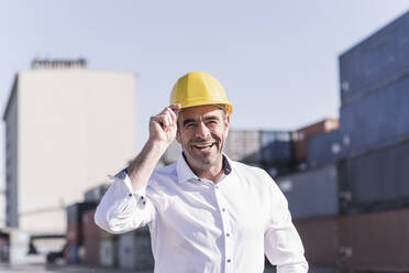 Porträt eines lachenden Geschäftsmannes mit Schutzhelm auf einem Industriegelände - UUF20417