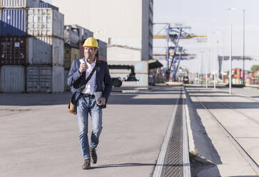 Businessman with smartphone and digital tablet walking in front of cargo containers - UUF20422