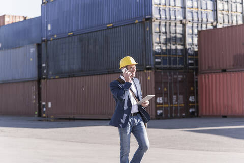 Geschäftsmann am Telefon mit digitalem Tablet vor Frachtcontainern gehend, lizenzfreies Stockfoto