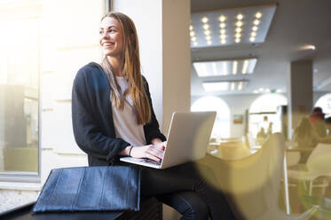 Happy woman looking through window while using laptop in coffee shop - DIGF12496