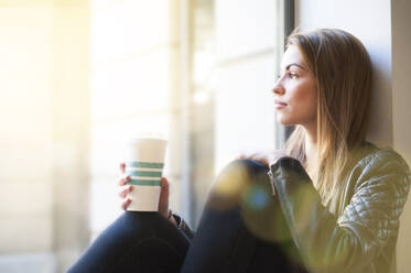 Frau, die durch ein Fenster schaut und einen Einweg-Kaffeebecher in einem Café hält - DIGF12499