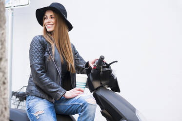 Glückliche junge Frau sitzt auf Motorroller gegen weiße Wand in der Stadt - DIGF12510