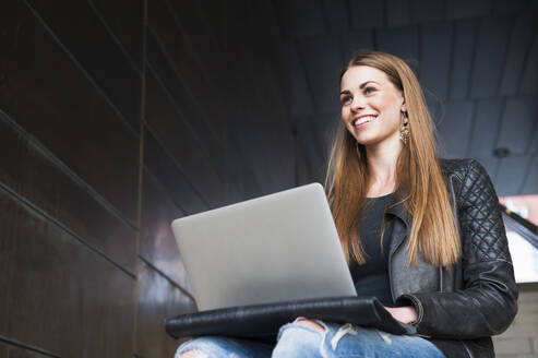 Lächelnde junge Frau, die einen Laptop benutzt, während sie in einer unterirdischen Gangway sitzt - DIGF12525