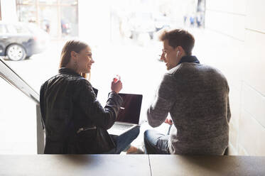 Young couple talking while sitting with laptop on steps in underground walkway - DIGF12528