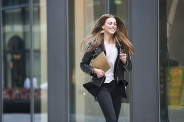 Fröhliche junge Frau mit Laptop beim Laufen in der Stadt - DIGF12531