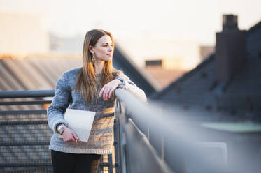Nachdenkliche junge Frau, die einen Laptop hält, während sie auf einer Gebäudeterrasse in der Stadt steht - DIGF12546