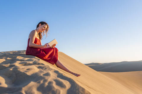 Frau in rotem Kleid sitzt in den Dünen und benutzt ein Tablet, Gran Canaria, Spanien, lizenzfreies Stockfoto