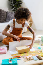 Smiling young woman sitting on the floor at home wrapping gifts - GIOF08256