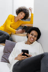 Porträt einer glücklichen jungen Frau, die sich mit ihrem Smartphone auf der Couch entspannt, während ihr Freund im Hintergrund steht - GIOF08273