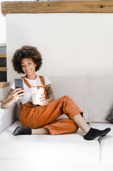 Porträt einer lächelnden jungen Frau, die zu Hause auf der Couch sitzt und eine Schachtel mit asiatischem Essen auf ihr Handy schaut - GIOF08290