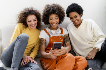 Gruppenbild von drei lachenden Freunden, die auf einer Couch sitzen und ein Selfie mit einem Smartphone machen - GIOF08304