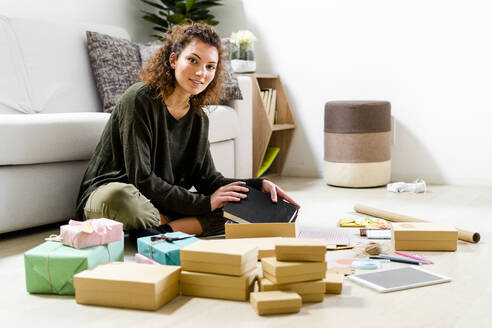 Portrait of smiling young woman sitting on the floor at home wrapping gifts - GIOF08316