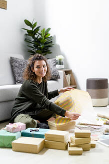 Portrait of smiling young woman sitting on the floor at home wrapping gifts - GIOF08318