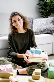 Portrait of smiling young woman sitting on the floor at home with wrapped presents - GIOF08324