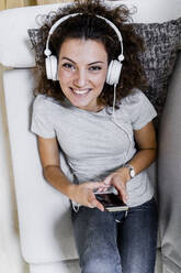 Portrait of smiling young woman lying on the couch listening music with headphones and smartphone - GIOF08332