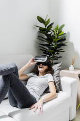 Junge Frau liegt zu Hause auf der Couch und benutzt eine Virtual-Reality-Brille - GIOF08336