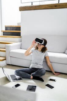 Junge Frau sitzt zu Hause auf dem Boden und benutzt eine Virtual-Reality-Brille und verschiedene elektronische Geräte - GIOF08337