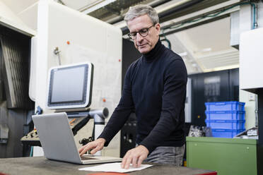 Männlicher Vorgesetzter, der ein Dokument liest, während er einen Laptop in einer Fabrik benutzt - DIGF12622