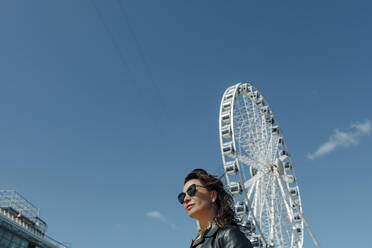 Frau mit Sonnenbrille in der Nähe des Riesenrads vor blauem Himmel - OGF00432