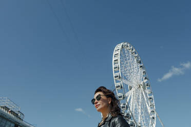 Woman wearing sunglasses near Ferris wheel against blue sky - OGF00432