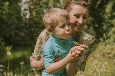 Glückliche Frau mit Sohn, der an einem sonnigen Tag Löwenzahnsamen im Garten ausbläst - MFF05851