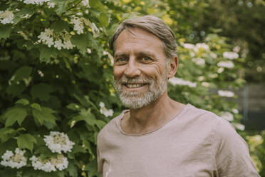 Smiling mature bearded man standing by flowering plant at garden - MFF05853