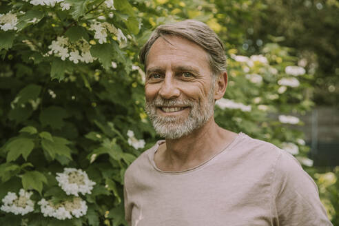 Lächelnder reifer bärtiger Mann, der an einer blühenden Pflanze im Garten steht - MFF05853