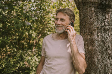 Smiling mature man talking on mobile phone while leaning on tree at garden - MFF05860