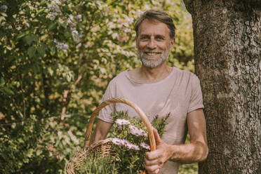 Happy bearded man holding basket with flowers and herbs while leaning on tree trunk at garden - MFF05862