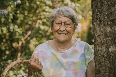 Glückliche ältere Frau im Ruhestand, die einen Korb hält, während sie an einem Baum im Garten steht - MFF05865