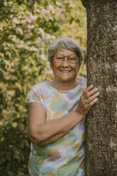 Glückliche ältere Frau im Ruhestand, die an einem Baum vor Pflanzen im Garten steht - MFF05867