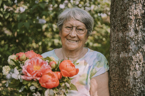 Retired senior woman holding fresh peony bouquet by tree at garden stock photo