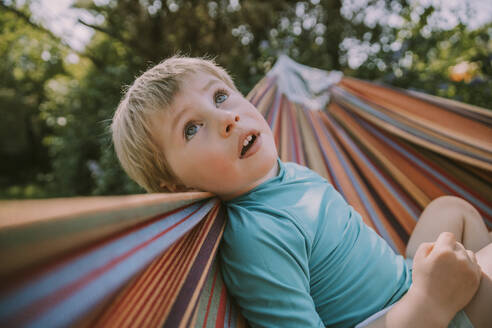 Niedlicher Junge schaut nach oben, während er sich in der Hängematte im Garten an einem sonnigen Tag entspannt - MFF05872