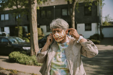 Retired woman wearing protective face mask while standing outdoors on sunny day - MFF05881