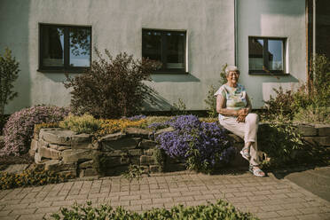 Lächelnde ältere Frau, die an einem sonnigen Tag am Haus sitzt - MFF05882