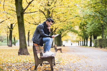 Junger Mann schaut auf ein digitales Tablet, während er auf einer Bank im Park sitzt - DIGF12637