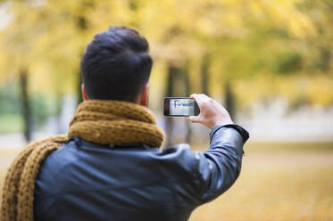 Young man photographing while standing at park during autumn - DIGF12641