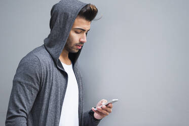 Handsome young man wearing hood while using smart phone by wall - DIGF12649