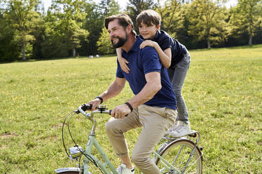 Lächelnder Vater und Sohn genießen eine Fahrradtour im Gras an einem sonnigen Tag - AUF00519